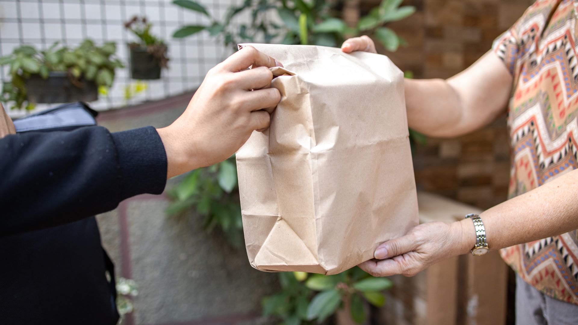 Two hands exchanging a brown paper bag in a plant-filled setting